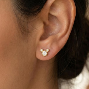 May include: Gold-colored stud earrings in the shape of a mouse head, with a large central stone and two smaller stones for ears. The earrings are set against the backdrop of a person's ear.