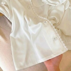 May include: White satin pajama set with delicate lace trim. The top features a button-down front, a chest pocket, and a scalloped lace collar. The matching shorts are visible in the background. The set is made of a soft, flowing fabric.