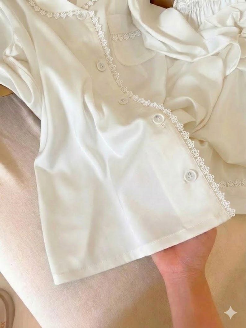 May include: White satin pajama set with delicate lace trim. The top features a button-down front, a chest pocket, and a scalloped lace collar. The matching shorts are visible in the background. The set is made of a soft, flowing fabric.