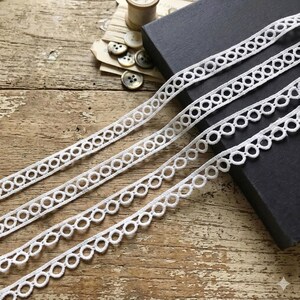 May include: Four strands of white lace ribbon with an open circle design are displayed diagonally across a wooden surface. The lace is next to a black box, buttons, and a spool of thread, suggesting a crafting or sewing theme.