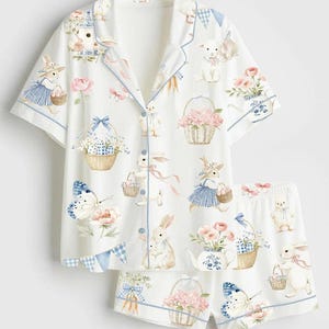 May include: White pajama set with a whimsical Easter-themed print. The set includes a short-sleeved button-up shirt and matching shorts. The print features bunnies, baskets of flowers, butterflies, and carrots, with blue trim.