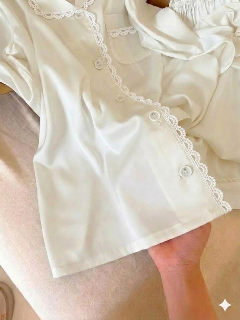 May include: White pajama set featuring a button-down top with a scalloped lace trim and a matching pair of shorts. The top has a classic collar and a chest pocket. The fabric appears to be a soft, lightweight material.
