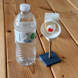 May include: A clear plastic water bottle with a white cap next to a white and red speaker on a stand. The speaker has a red logo and is mounted on a blue base. The items are on a wooden surface.
