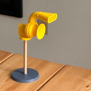 May include: A yellow, stylized microphone sculpture with a round base and a wooden stem. The microphone has a circular body and a curved, horn-like top. The base is a muted blue color, and the sculpture is set on a wooden surface.
