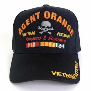 May include: Black baseball cap with embroidered text and graphics. The cap features the words "AGENT ORANGE" in orange, with "VIETNAM VETERAN" and "Sprayed & Betrayed" also embroidered. A skull and crossbones graphic is centered above a row of colorful ribbons.