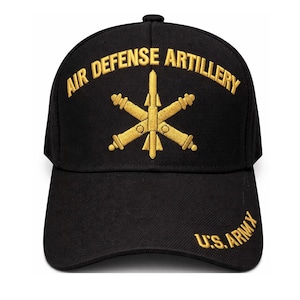 May include: Black baseball cap with gold embroidered text and emblem. The text reads "AIR DEFENSE ARTILLERY" and "U.S. ARMY". The emblem is a gold starburst design. A classic, adjustable hat.