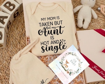 May include: A beige ribbed baby bodysuit with the black text MY MOM IS TAKEN BUT But my Aunt IS HOT AND SINGLE, a matching beige ribbed baby hat with the text hello world, and a square card featuring a floral wreath and a deer illustration.