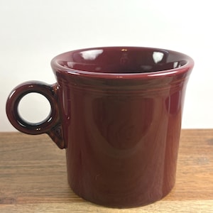 May include: A solid deep burgundy ceramic mug with a glossy finish. The mug features a wide, cylindrical body and a circular handle attached to the side. The rim is slightly flared, and the overall design is simple with no patterns or text.