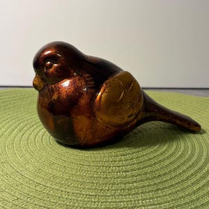 May include: A decorative bird figurine in shades of brown and gold. The bird has a rounded body, a small beak, and a textured wing detail. The figurine is placed on a green, circular placemat.