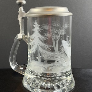 May include: A clear glass beer stein featuring an etched forest scene with a deer and pine trees. The stein has a sturdy glass handle and a hinged pewter lid with a decorative finial. The base of the glass is thick and has a radial pattern.