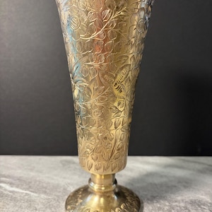 May include: A gold-colored vase with an intricate floral pattern. The vase has a flared top, a pedestal base, and is approximately 8 inches tall. The detailed design covers the entire surface, creating a textured appearance.