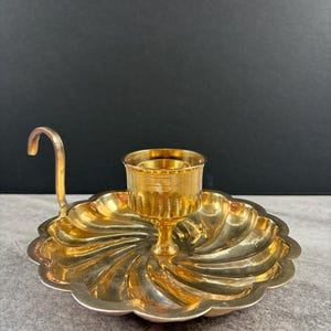 May include: A gold-coloured candle holder with a scalloped, spiral-patterned base and a small handle. The candle holder has a cup-shaped centre to hold a candle. The metal has a shiny finish.
