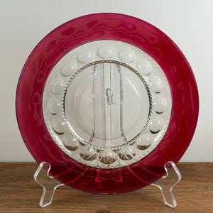May include: A circular glass plate featuring a deep red outer rim and a clear center section. The clear portion is decorated with a ring of raised, rounded glass bubbles and a textured, scalloped inner border surrounding a smooth, transparent base.