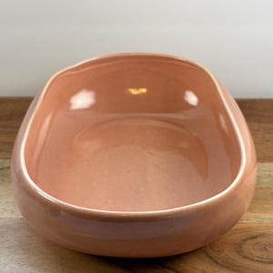 May include: An oval-shaped ceramic bowl with a smooth, glossy finish in a soft peach color. The bowl features a deep, rounded basin with thick, curved walls and a continuous, uniform rim, presenting a simple and functional design for serving.