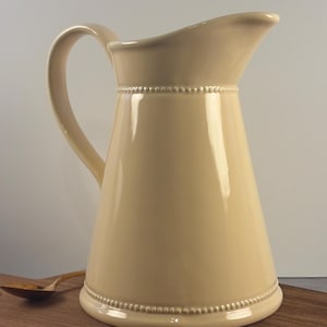May include: Cream-colored ceramic pitcher with a curved handle and a wide spout. The pitcher has a beaded design around the base and the top rim. A wooden spoon rests on a wooden surface next to the pitcher.