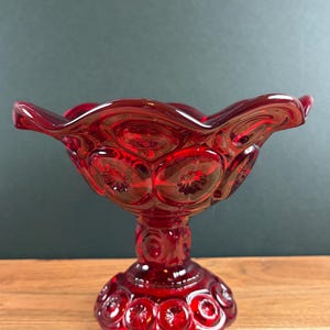 May include: A ruby red glass compote dish featuring a pressed moon and star pattern. The bowl has a wide, scalloped rim and sits on a pedestal base. The glass is translucent and deep red, with circular indentations containing star motifs.