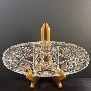 May include: An oval clear glass dish featuring intricate cut patterns including starburst motifs, geometric cross-hatching, and a central hexagonal design. The dish has a scalloped edge and is displayed on a gold-colored stand.