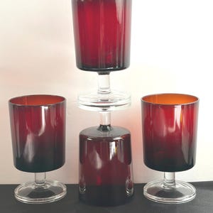 May include: A set of four deep red glass goblets with clear glass stems and circular bases. Two goblets stand upright on the left and right, while the other two are stacked vertically in the center, with one upright and one inverted on top.