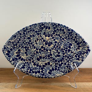 May include: An oval-shaped ceramic platter featuring a dense, repeating pattern of dark blue and white teardrop-shaped motifs. The dish is displayed upright on a clear acrylic stand, showcasing the intricate, hand-painted style of the blue and white glaze.
