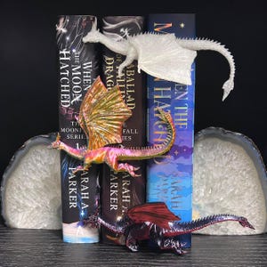 May include: Three dragon figurines displayed with books. One is white and glittery, one is iridescent gold and pink, and one is dark purple with red wings. The books are titled When the Moon Hatched and A Ballad of Fallen Dragons by Sarah A. Parker.