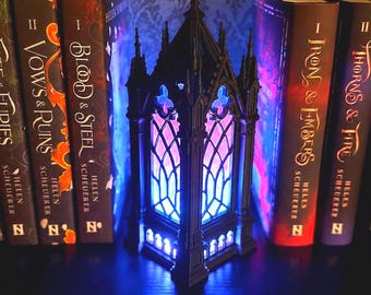 May include: A black, 3D-printed gothic cathedral-style lamp with pointed arches and intricate tracery, illuminated from within by a vibrant blue and purple light, positioned between several fantasy book spines on a dark surface.