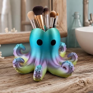 Puede incluir: Un soporte para brochas de maquillaje con forma de pulpo que presenta un degradado de colores que va del azul verdoso en la parte superior al morado y verde lima en la base. El soporte tiene dos ojos negros circulares y contiene brochas.