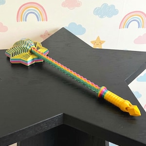May include: A multi-colored, 3D-printed star-shaped wand featuring a long, wavy, rainbow-gradient shaft and a solid yellow handle with a pointed pommel. The star head displays a layered, concentric design with vibrant stripes of pink, blue, and green.