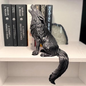 May include: A matte black wolf figurine in a howling pose with white highlights on its fur. The wolf is seated with its long, textured tail hanging over the edge of a white shelf, positioned in front of a row of books with black and white spines.