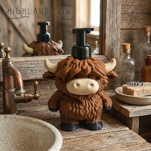 May include: A brown Highland Cow soap dispenser with a black pump top, white horns, and a beige snout. The dispenser is on a wooden surface with the text "HIGHLAND COW" above it. The dispenser is in a bathroom setting.