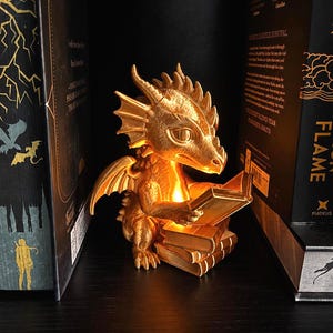 May include: A golden dragon figurine with a textured surface, illuminated from within, sitting on a stack of books while holding an open book. The dragon features detailed wings, horns, and scales, casting a warm glow on the surrounding pages.