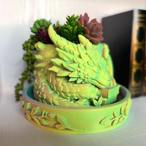 May include: A lime green and yellow dragon-shaped planter resting on a matching circular tray with leaf relief details. The dragon is curled around a small book, and the planter contains several artificial green and red succulents.