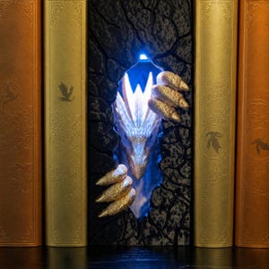 May include: A book nook insert featuring a textured black stone wall with a jagged opening. A glowing white dragon head emerges from the center, flanked by two golden, clawed dragon feet. The insert is positioned between two gold-colored books.