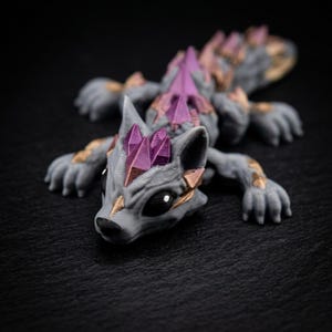 Puede incluir: Una criatura de fantasía gris, impresa en 3D, con cabeza de lobo y cuerpo de lagarto. Presenta detalles de cristales morados en la cabeza y la espalda, con detalles color bronce en el cuerpo. La criatura está tumbada sobre una superficie oscura.