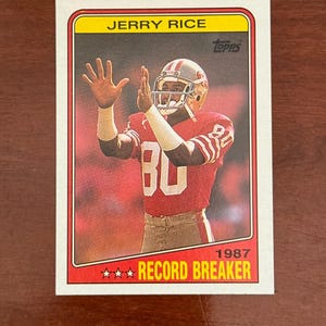 May include: A 1987 Topps Jerry Rice football card featuring the player in a red San Francisco 49ers jersey with the number 80, wearing a helmet and white arm sleeves, with the text JERRY RICE at the top and RECORD BREAKER at the bottom.