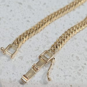 May include: Close-up of a gold curb chain bracelet with an open rectangular clasp. The bracelet's links are interlocked, and the clasp is open, revealing the intricate design. The bracelet is displayed on a light-coloured surface.