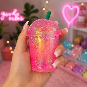 Könnte beinhalten: Ein transparenter Kunststoffbecher gefüllt mit leuchtend rosa Flüssigkeit und irisierendem Glitzer, ausgestattet mit einem passenden rosa Kuppeldeckel und einem einfarbigen grünen Strohhalm. Der Becher hat ein strukturiertes, glitzerndes Aussehen.