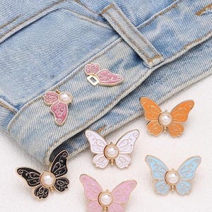 May include: A collection of seven butterfly-shaped pins featuring enamel wings in black, white, orange, light blue, and various shades of pink. Each butterfly pin is centred with a small, round, white faux pearl and finished with gold-tone metal.