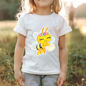 K&ouml;nnte beinhalten: Wei&szlig;es T-Shirt mit einem Cartoon-Bienen-Design. Die Biene ist gelb, schwarz und wei&szlig;, tr&auml;gt einen Blumenkranz und einen Korb. Das T-Shirt wird von einem Kind getragen.