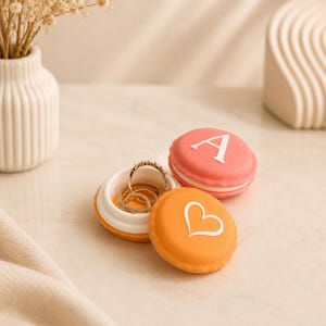 Könnte beinhalten: Eine kleine, runde Schmuckdose in Macaron-Form in Korallenrosa mit einem weißen Buchstaben A auf dem Deckel, dazu ein orangefarbener Deckel mit weißem Herzumriss. Die offene Basis enthält mehrere dünne Ringe aus goldfarbenem Metall im Inneren.