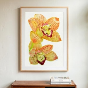 Puede incluir: Una impresión en acuarela enmarcada de dos flores de orquídeas de color naranja y amarillo con detalles en rojo y verde. La obra de arte se exhibe en una pared blanca, encima de un mueble de madera. El marco es de color marrón claro.