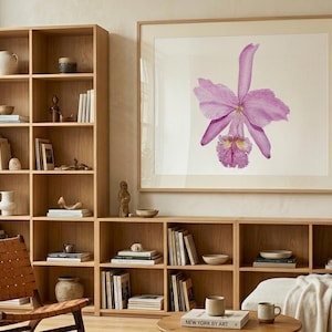 Puede incluir: Un salón con estanterías de madera clara, una silla de cuero y una gran impresión enmarcada de una orquídea púrpura. La habitación tiene una paleta de colores neutros y una estética moderna. Libros y objetos decorativos se exhiben en las estanterías.