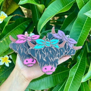 May include: Three Highland cow ornaments with black fur, pink noses, and colourful bows. The bows are pink, teal, and purple. The ornaments are held in a hand against a background of green leaves and flowers.