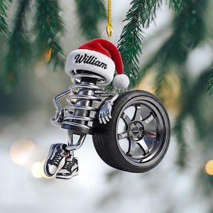 May include: A hanging ornament featuring a metallic coil spring character wearing a red and white Santa hat with the name William printed on the brim. The character has metallic arms and legs, wears black and white sneakers, and leans against a dark gray car wheel.