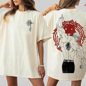 May include: Cream-colored t-shirt featuring a black and white graphic of a weapon on the front and a muscular figure with a lion emblem on the back. The shirt has short sleeves and a relaxed fit, suitable for casual wear.