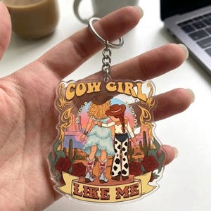 May include: A clear acrylic keychain featuring a colorful illustration of two figures in western attire standing in a desert landscape with cacti and red roses. The text COW GIRL is printed at the top and LIKE ME is printed on a banner at the bottom.