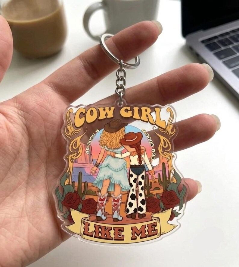 May include: A clear acrylic keychain featuring a colorful illustration of two figures in western attire standing in a desert landscape with cacti and red roses. The text COW GIRL is printed at the top and LIKE ME is printed on a banner at the bottom.