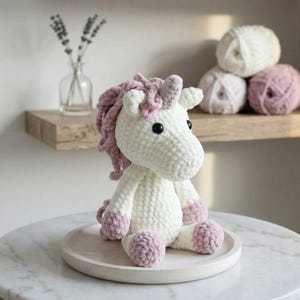 May include: A handmade white crochet unicorn with pink mane, horn, and hooves. The plush toy has black eyes and is sitting on a wooden tray. The background includes a shelf with yarn balls and a vase with lavender.