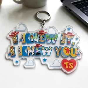 May include: A clear acrylic keyring featuring the text I KNEW IT, I KNEW YOU in blue block letters. The letters incorporate patterns inspired by cowboy and space ranger characters, including hats, stars, and clouds. A red heart with TS is included.