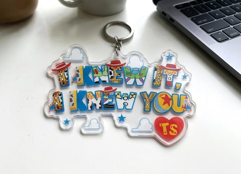 May include: A clear acrylic keyring featuring the text I KNEW IT, I KNEW YOU in blue block letters. The letters incorporate patterns inspired by cowboy and space ranger characters, including hats, stars, and clouds. A red heart with TS is included.