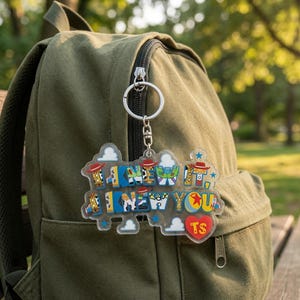 May include: A clear acrylic keyring featuring the text I KNEW IT, I KNEW YOU in blue, yellow, and white block letters. The design includes small red cowboy hats, white clouds, a yellow star, and a red heart with the letters TS inside.